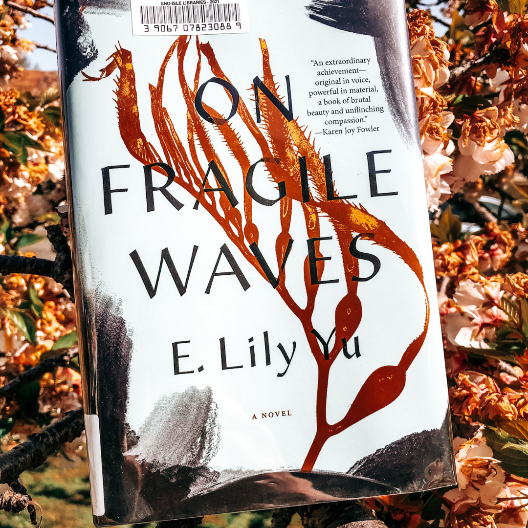 [ID: The book On Fragile Waves by E. Lily Yu sits in a cherry tree.]